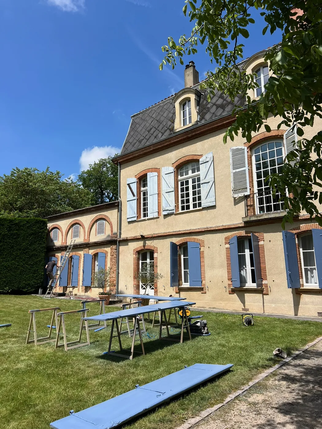 Chantier de rénovation : volets en bois en cours de peinture sur tréteaux devant la maison de maître, ManufactureVB à l'ouvrage