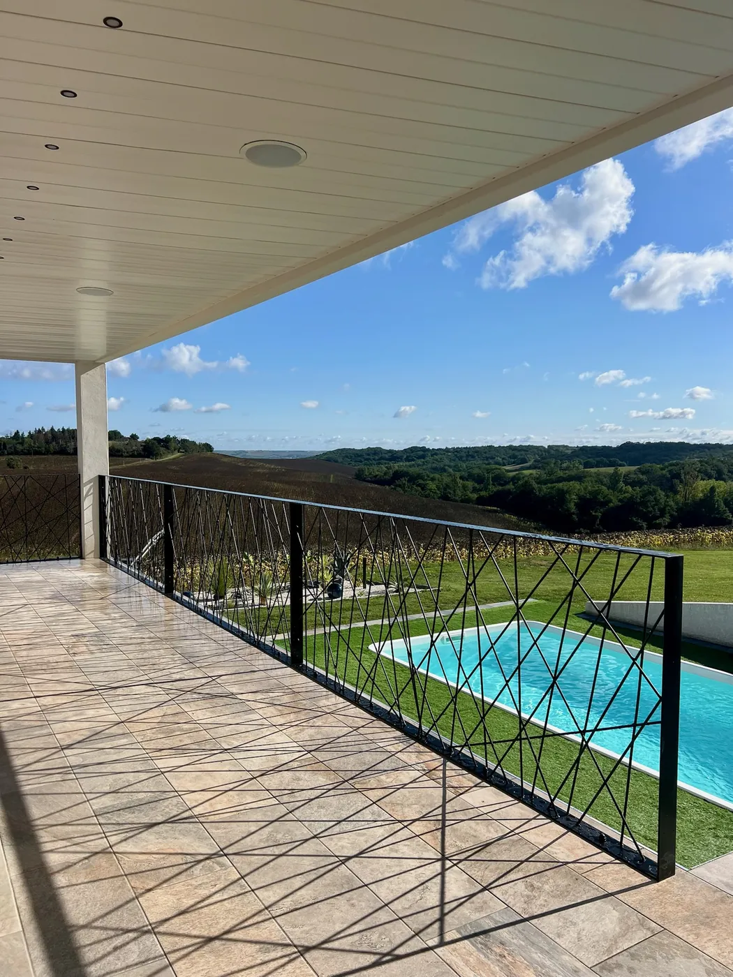 Garde-corps en acier thermolaqué noir à motifs croisés bordant une terrasse couverte avec vue sur la piscine et la campagne, réalisation ManufactureVB